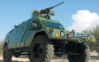 A green Light Armored Vehicle with large tires and a mounted machine gun on top is parked outdoors under a blue sky.