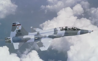 A military jet with a gray and blue camouflage pattern flies above the clouds, viewed from the side. The cockpit canopy is open and the aircraft markings are visible on the tail and fuselage.