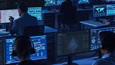 People wearing headsets sit at computer desks in a dark room, monitoring multiple large screens displaying data, maps, and graphics, suggesting a high-tech operations or control center environment.