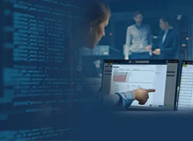 A person points at a computer screen displaying data, with lines of code visible on the left. In the background, two people are standing and talking in a dimly lit office setting.