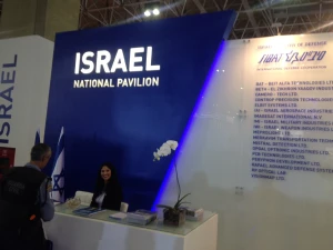 A woman sits at a reception desk in front of a blue and white booth labeled Israel National Pavilion, with a list of organizations displayed on the wall and an orchid plant on the counter.