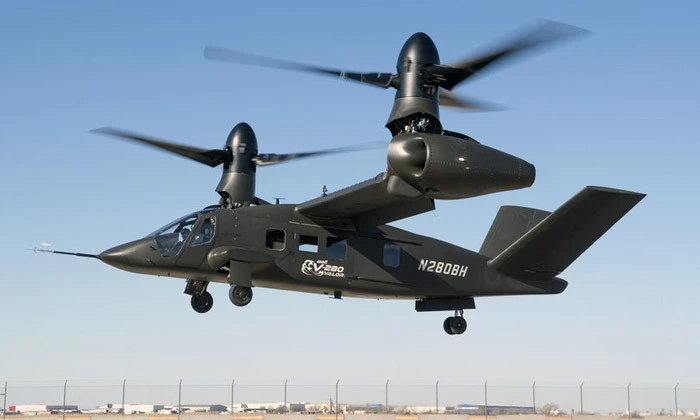 A black tiltrotor aircraft with two large propellers is flying above a concrete surface on a clear day. Featuring advanced helicopter seating systems, the V-280 Valor (N280BH) is seen with buildings and a fence in the background.