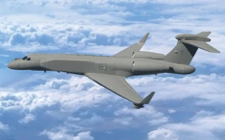 A grey military surveillance aircraft with a radar pod on its tail is flying above the clouds during the day, viewed from the side against a blue sky.