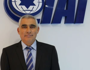A man in a suit and striped tie stands in front of a wall featuring the blue IAI logo and a symbol with a star inside a circle.