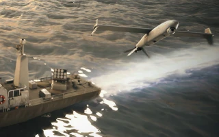 A military drone flies low over the ocean next to a naval ship, creating a white trail on the water’s surface as the sun reflects off the waves.