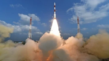A rocket carrying a nanosatellite launches into the sky, surrounded by clouds of smoke and flanked by tall launch towers, against a backdrop of blue sky and scattered clouds.