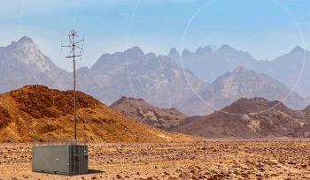 A metal shipping container with an antenna tower stands in a rocky, barren desert landscape with rugged mountains in the background under a clear blue sky.