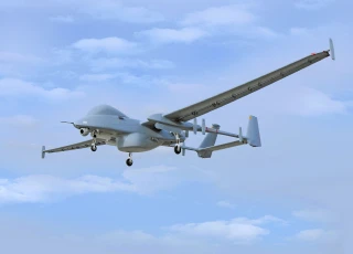 A large gray unmanned aerial vehicle (drone) with long wings is flying in the sky with landing gear extended, against a background of light clouds.