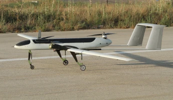 A medium-sized, twin-boom unmanned aerial vehicle (drone) with a black and white body, often seen in Indian aviation, is parked on a paved surface, with grass and plants in the background.