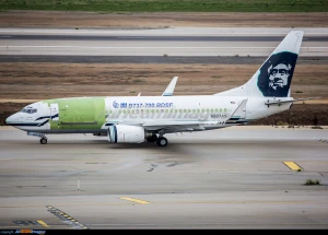 A green and white Alaska Airlines plane, possibly undergoing Freighter Conversion, taxis on the concrete runway. The aircraft features a large bearded face on its tail, with grass and runway markings visible in the background.