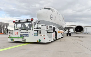 A white Lufthansa airplane is being towed on an airport runway by a green and white electric tow tractor labeled “TaxiBot.” The sky above is cloudy and airport buildings are visible in the background.