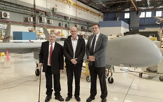 Three men in suits stand in front of a large, gray unmanned aerial vehicle (drone) inside a spacious industrial facility, highlighting the role of blockchain security amid equipment, tools, and machinery in the background.