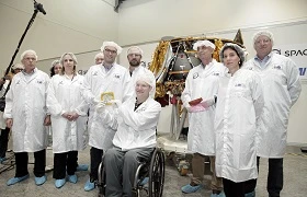 A group of people in white lab coats, hair covers, and blue shoe covers pose indoors near a spacecraft model. Highlighting advancements like blockchain security, one person in a wheelchair holds a small object and smiles at the camera.