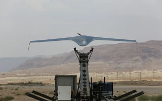 A military drone with a wide wingspan is mounted on a launcher, ready for takeoff on a flat, open area with mountains and cloudy sky in the background.