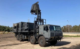 A large military vehicle with a raised radar system on top is parked on a dirt lot under a clear blue sky. The vehicle is painted in dark colors and has rugged, off-road tires.