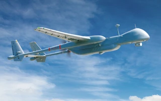 A large, gray unmanned aerial vehicle (UAV) flies in a clear blue sky with scattered clouds. The drone has long wings, a prominent nose, and a tail fin, appearing streamlined and modern.