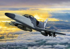 A military jet aircraft flying above clouds at sunset, with the sky glowing orange and yellow in the background. The jet is gray with visible markings and weaponry attached under its wings.