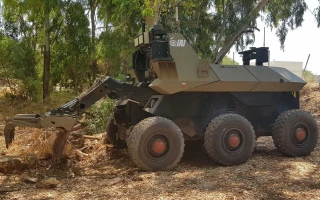 A military robot vehicle with six large wheels and a robotic arm is operating outdoors on dry, uneven terrain with trees in the background.