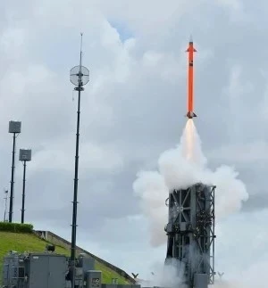 A bright orange missile launches vertically from a military platform, leaving a trail of smoke against a cloudy sky, with equipment and antennas visible in the foreground.