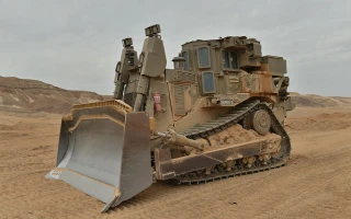A large armored bulldozer with reinforced windows and heavy-duty treads is parked on sandy terrain under a cloudy sky.