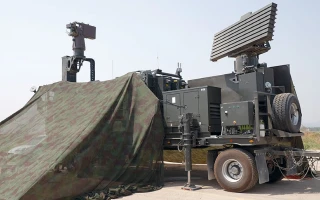 A military radar system mounted on a trailer is partially covered by camouflage netting, with two large radar panels and various equipment visible under a clear sky.
