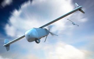 Two unmanned aerial drones are flying in the sky with clouds in the background. The larger drone is in the foreground and the smaller one is farther in the distance.
