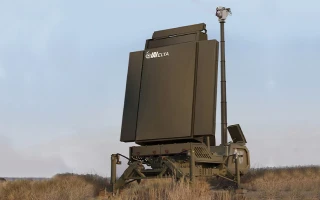 A large military radar system with the label EL/M-2084, part of advanced air defence systems, is positioned on a grassy field, mounted on a movable platform with a tall mast and sensor unit extending upward.