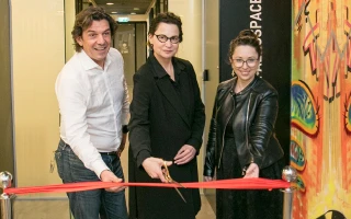 Three adults stand in front of a red ribbon at an indoor event. The woman in the center, holding scissors, prepares to cut the ribbon at the new Innovation Center, while the two others hold the ribbon on either side.