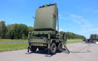 A large green military radar system mounted on the back of a truck is positioned on a paved road, with stabilizing legs extended. Trees and open sky are visible in the background.