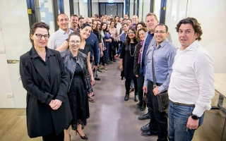 A large group of people, dressed in business casual attire, stands in two lines facing each other in an office hallway, smiling at the camera.