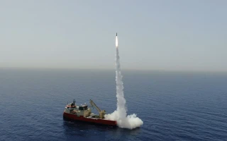 A missile firing launches vertically from a platform on a ship in the middle of the ocean, leaving a trail of white smoke against a clear sky and calm water.