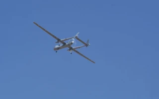 A German UAV twin-engine propeller aircraft flies against a clear blue sky, viewed from below.