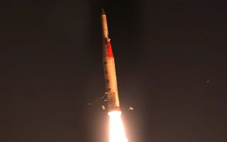 A rocket launches into the night sky, with bright flames and smoke trailing from its base, illuminating the dark background—reminiscent of Israel's air defence intercepting threats overhead.