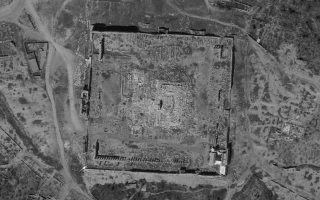 Aerial view of ancient temple ruins with a large, nearly square layout, surrounded by crumbling stone walls and scattered remnants within the structure, set in a sparsely vegetated landscape.
