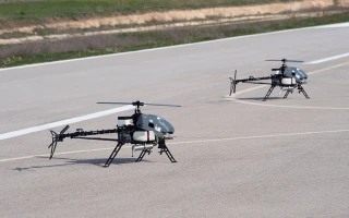 Two small unmanned helicopters with mounted equipment are positioned on a paved surface, possibly a runway, with grassy areas in the background.