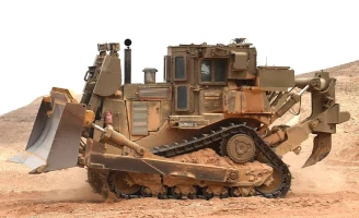 A large, armored bulldozer with heavy treads and a reinforced blade, designed for mining automation, moves earth in a sandy, desert-like environment. The machine is painted in a camouflage pattern.