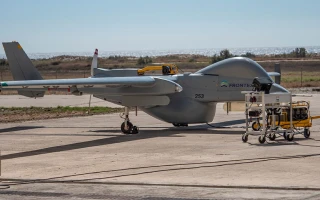 A large gray Frontex drone is parked on a tarmac next to ground support equipment, with an open field and distant water or coastline in the background.