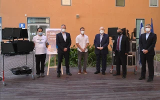 Six people wearing face masks stand in a row outdoors near computer equipment on carts, in front of a building with orange walls and green plants.