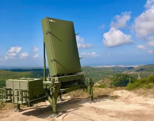 A large, green Radar Systems installation stands on a dirt path in a hilly landscape beneath a blue sky with scattered clouds. Distant hills and a body of water complete the scenic background.