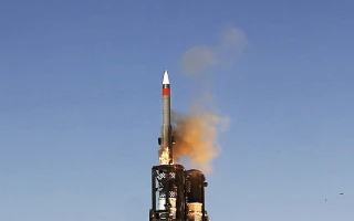 A missile is being launched vertically into a clear blue sky, with flames and smoke visible at its base, coming from a launch platform or launcher.