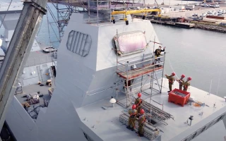 Five workers in protective gear stand on and around scaffolding set up against the superstructure of a naval ship docked at a port, performing maintenance or installation work.