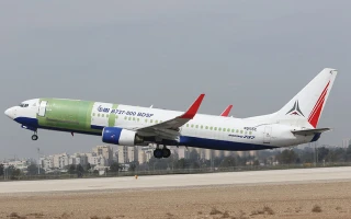 A commercial airplane with an unfinished green fuselage and red winglets, showcasing aircraft renovation solutions, takes off from a runway with a city skyline in the background.