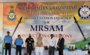 Five men stand on a stage in front of a banner reading “South Western Air Command Welcomes You to Air Force Station Jaisalmer for MRSAM Induction Ceremony,” flanked by two large images of missiles.