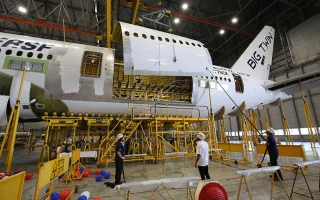 Workers stand on platforms inspecting and repairing the fuselage of a large airplane inside a hangar, with the tail labeled BIG THAI. The airplanes side panels are open, exposing the interior structure.