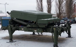 A large green military radar system with four sturdy legs is set up on a snowy outdoor surface, with trees, parked cars, and buildings visible in the background.
