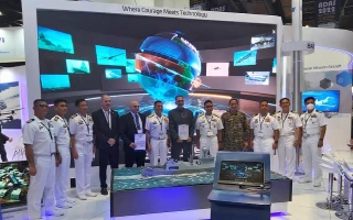 A group of naval officers and officials pose in front of a large digital display showing a 3D globe and aircraft images at a technology exhibition booth. Signs above read, “Where Change Meets Technology.”.