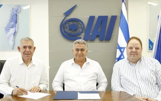 Three men in white shirts sit at a table with financial documents, in front of an IAI logo and Israeli flags on the wall behind them.