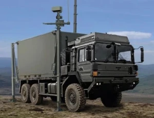 A military truck with large off-road tires is parked on rough terrain. Mounted on the truck is a tall mast with comjam and radar equipment. The background shows an open landscape and cloudy sky.