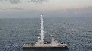 A naval warship in open water launches an advanced cruise missile vertically, leaving a trail of white smoke as it ascends into the sky. The sea appears calm under a cloudy sky.
