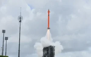 An orange rocket from Belbooth Aero launches vertically into a cloudy sky, leaving behind a trail of white smoke, with communication towers visible in the background.
